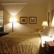 Het slaapgedeelte van de suite, met zicht op de badkamer. Creme-witte wanden met litho's en tekeningen, klassieke donkere meubels op een prachtige houten vloer, een twee meter breed bed geflankeerd door grote staande lampen die een gouden glans over het geheel laten schijnen.