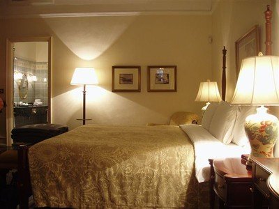 Het slaapgedeelte van de suite, met zicht op de badkamer. Creme-witte wanden met litho's en tekeningen, klassieke donkere meubels op een prachtige houten vloer, een twee meter breed bed geflankeerd door grote staande lampen die een gouden glans over het geheel laten schijnen.