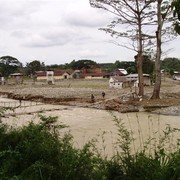 Bukit Lawang, een half jaar na de overstroming. Een kaal veld met enkele restanten van huizen en gebouwen. Voor ons een snelstromende rivier. Boven de kolkende geelbruine stroom hangt een smalle loopbrug.    