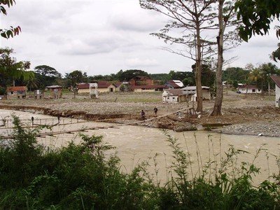 Bukit Lawang, een half jaar na de overstroming. Een kaal veld met enkele restanten van huizen en gebouwen. Voor ons een snelstromende rivier. Boven de kolkende geelbruine stroom hangt een smalle loopbrug.    
